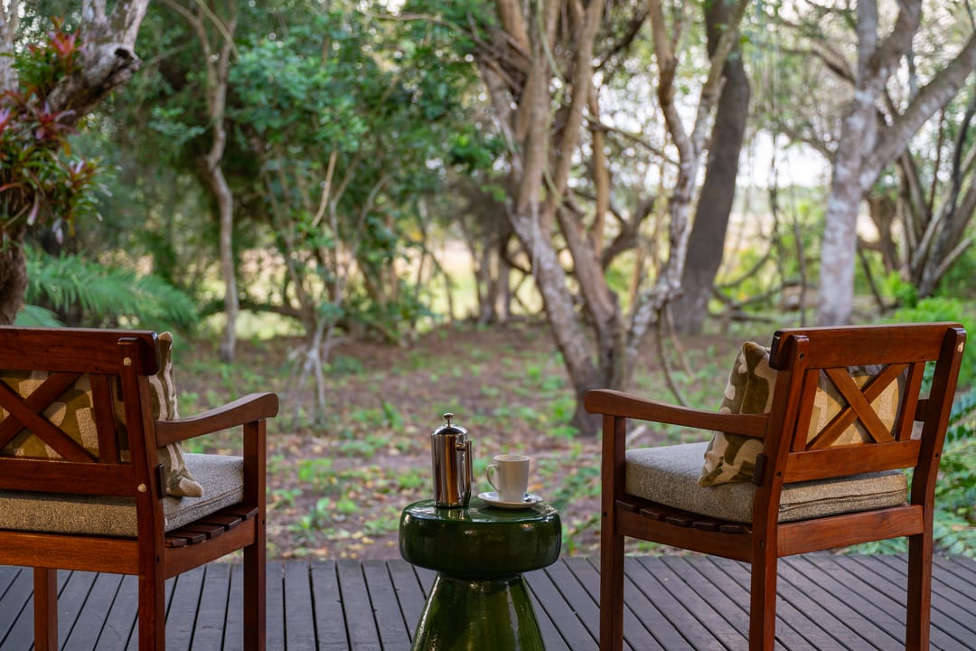 Two timber armchairs with cushions and a green side table on a suite deck, forest beyond.