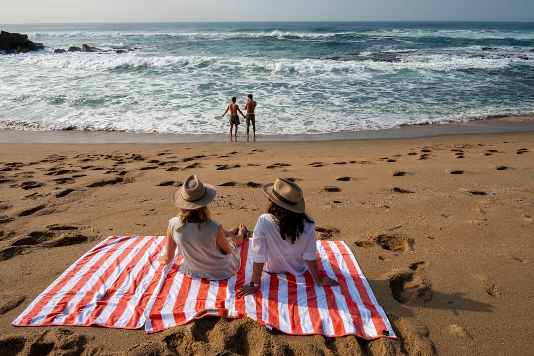 Two guests on a striped towel watch companions at the water’s edge across footprinted sand