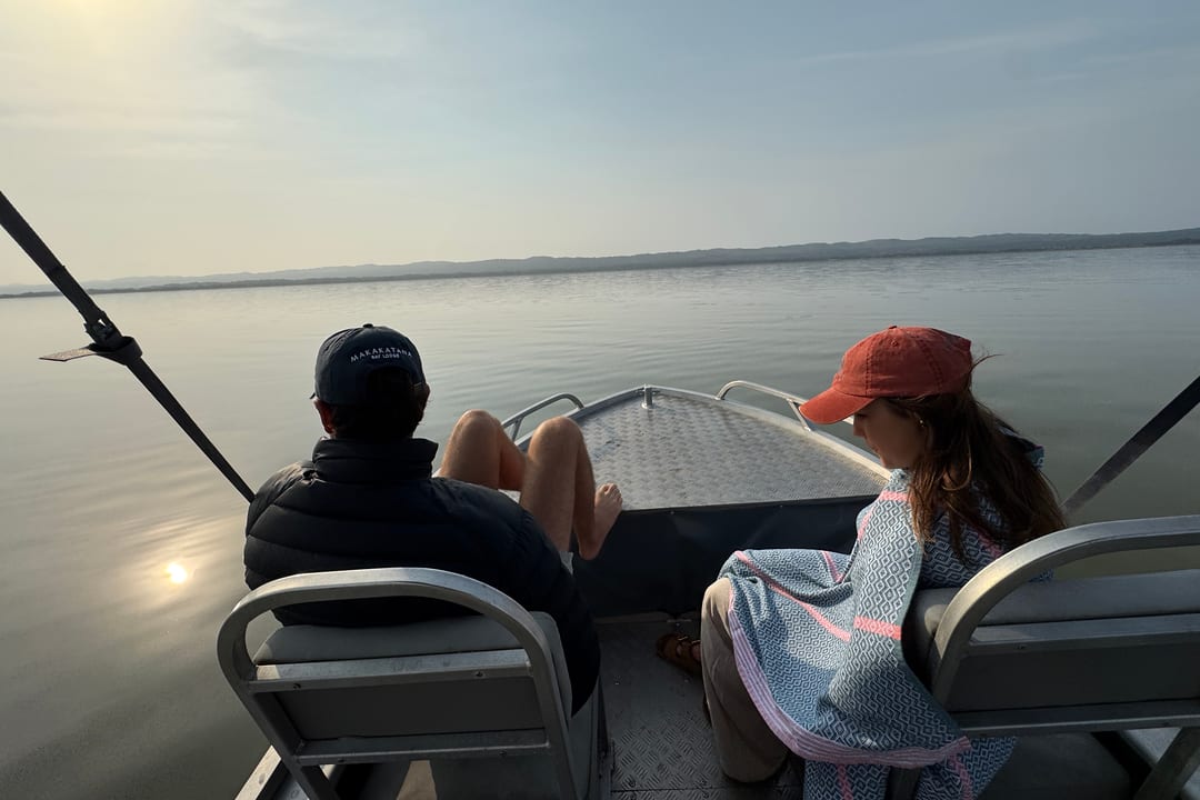 A couple sit at the bow as the boat crosses calm water towards a low sun