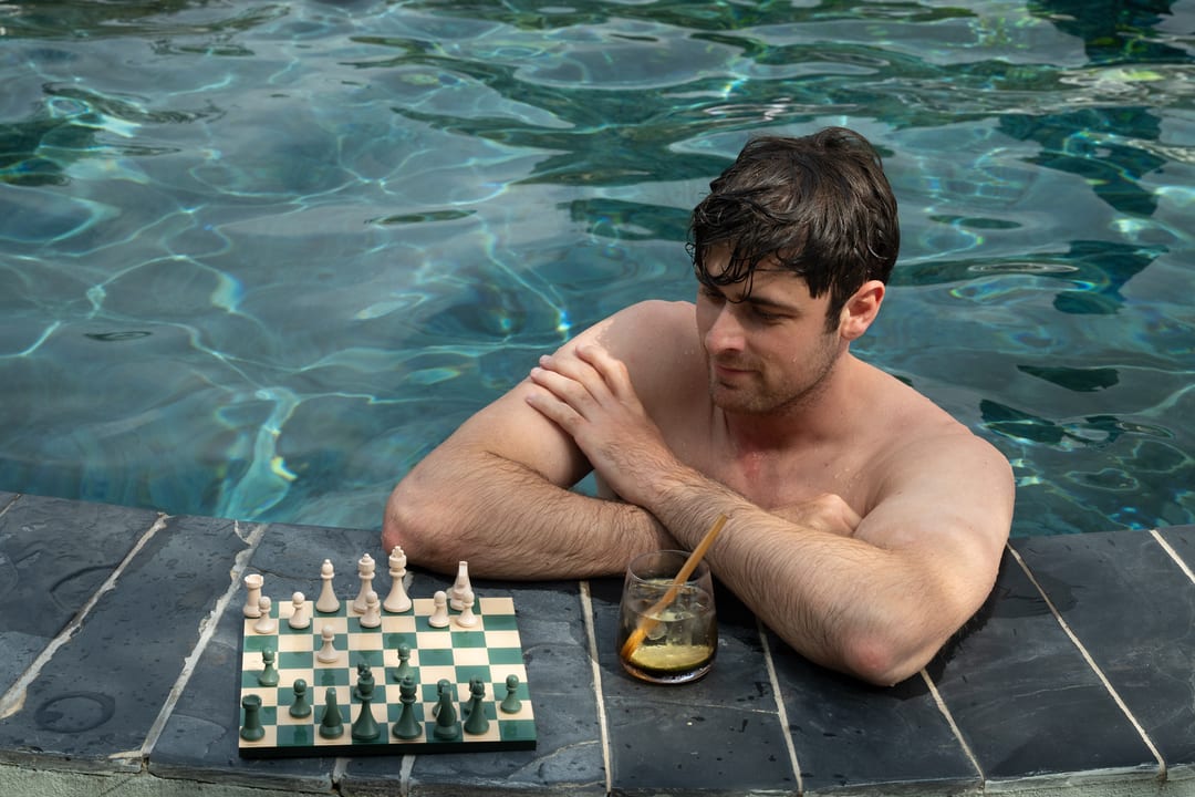 A guest leans on the pool edge beside a chess board and a cocktail