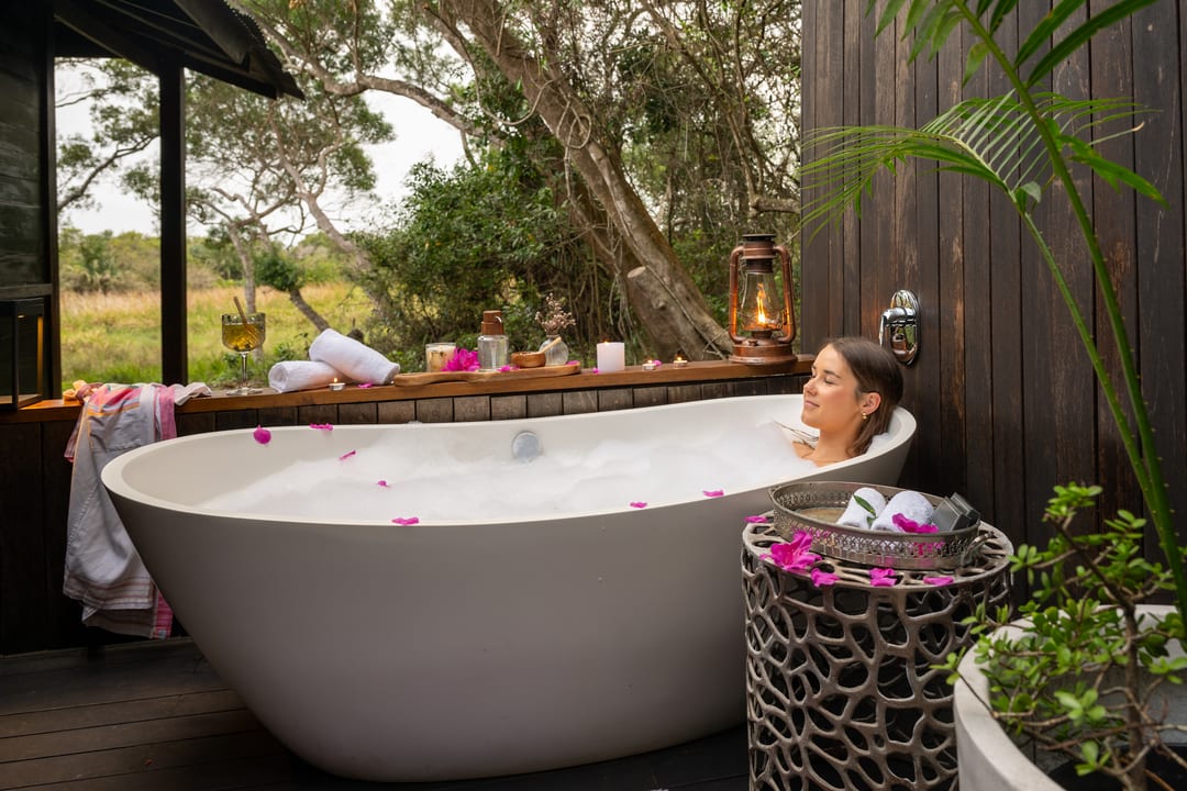 A guest relaxes in an outdoor bath scattered with petals, a lantern lit and the forest beyond