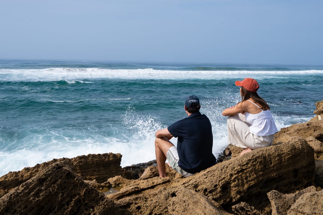 A couple sit on golden rocks watching the surf break below