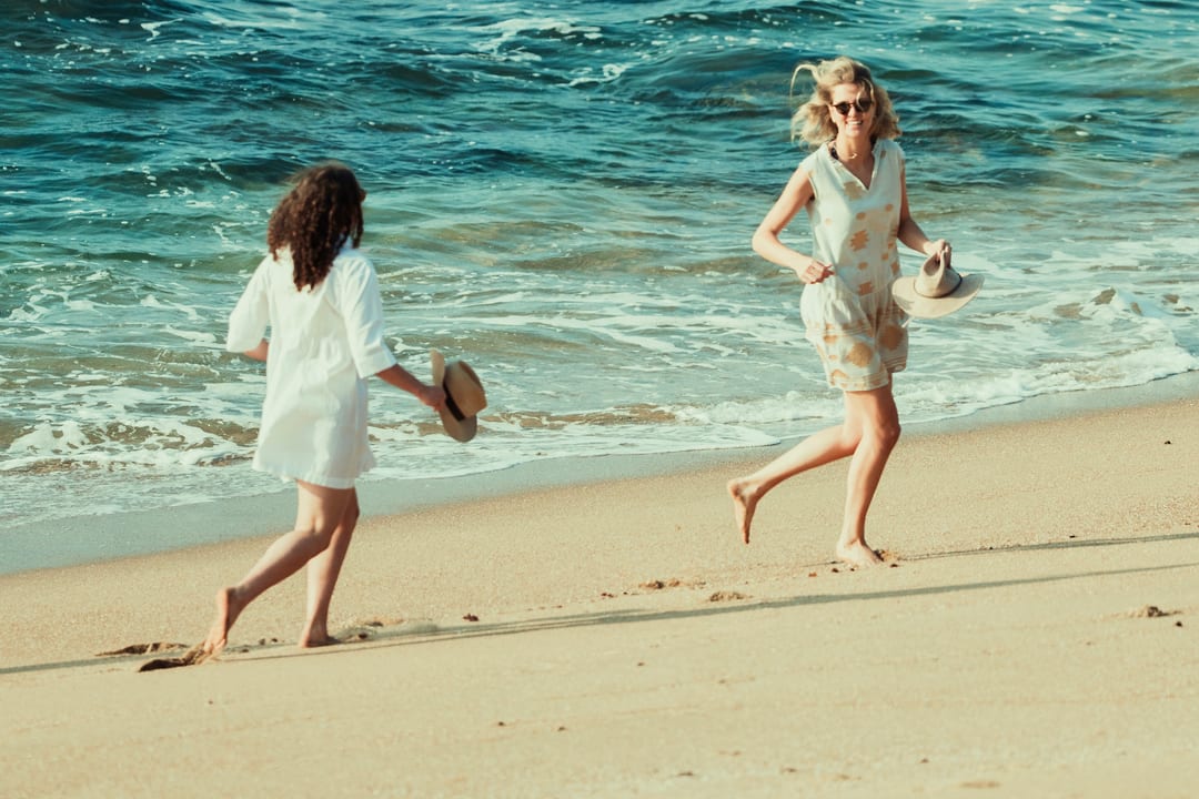 Two guests run barefoot along the shore swinging their sunhats, teal sea behind
