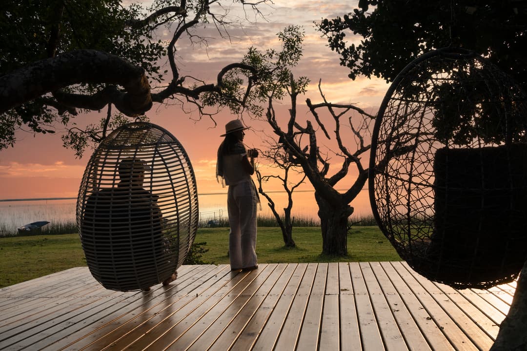 Sunrise on the waterside deck, a guest with a cup between two hanging chairs by the lake.