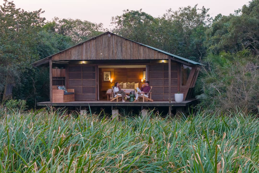 A lamp-lit suite on stilts seen from the reeds at dusk, two guests on its deck